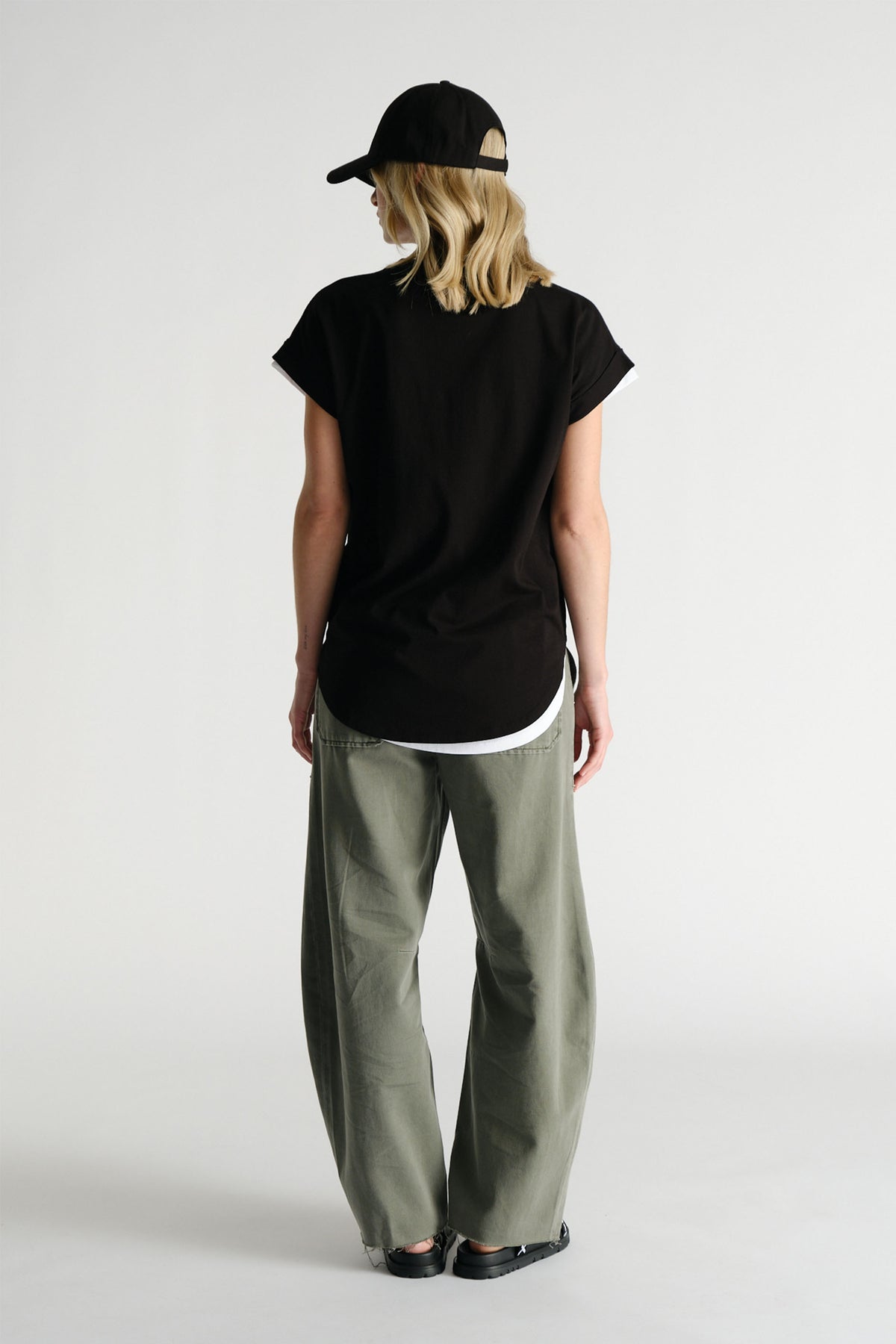 Rear view of woman wearing black D-Luxe Collegiate Tonal Tee with olive wide-leg pants and black cap by Decjuba Australian brand