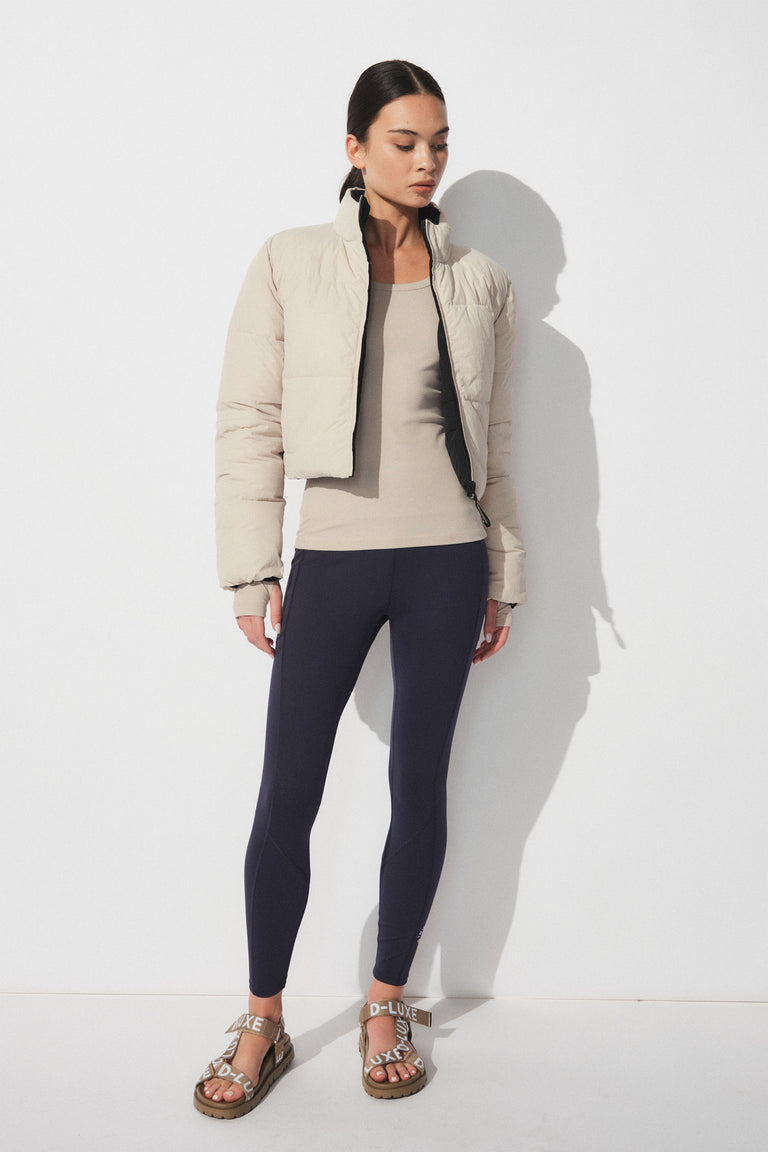Woman wearing a cropped beige puffer jacket over a neutral tank top, paired with navy slim-fit leggings and branded sandals