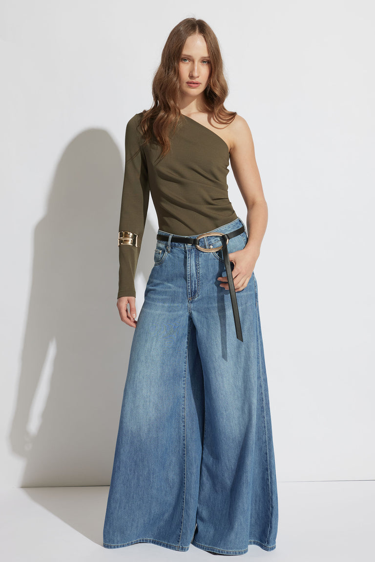 Woman wearing an olive one-shoulder top with a metallic cuff detail paired with wide-leg light blue jeans and a black belt.