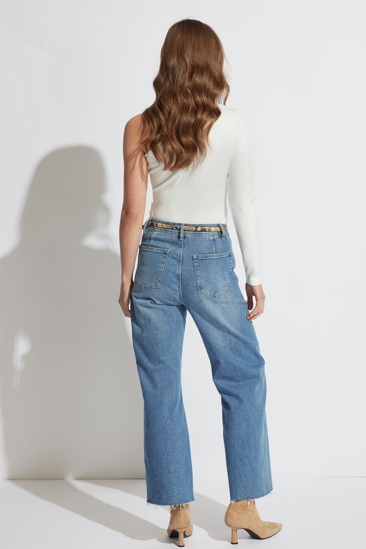 Woman with wavy hair wearing an ivory one-shoulder top paired with straight-leg blue jeans and tan ankle boots.