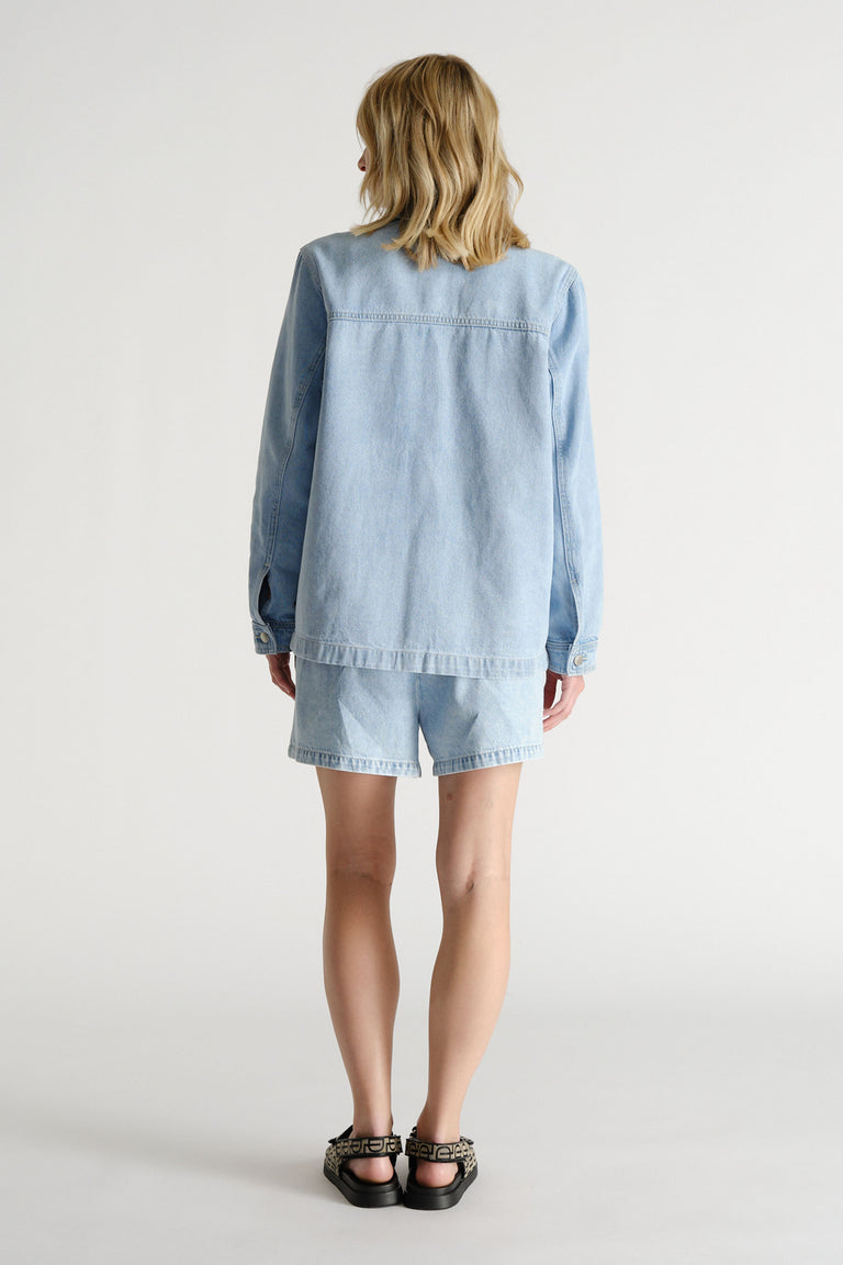 Woman seen from behind wearing light blue denim shacket and matching shorts with black sandals against a plain backdrop