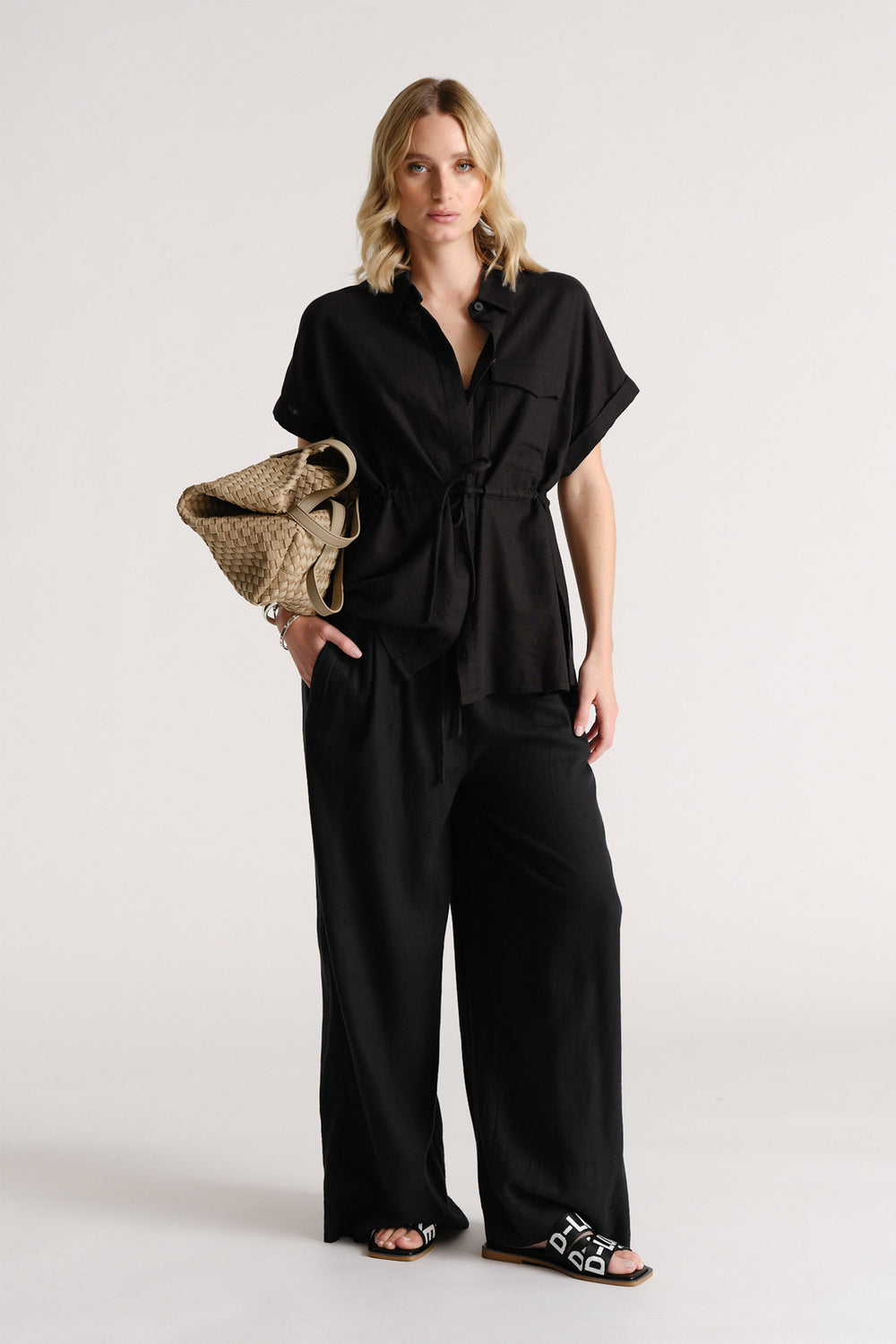 Woman wearing a relaxed black linen blend short-sleeve shirt with wide-leg black pants and black slide sandals.