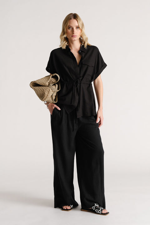 Woman wearing a relaxed black linen blend short-sleeve shirt with wide-leg black pants and black slide sandals.