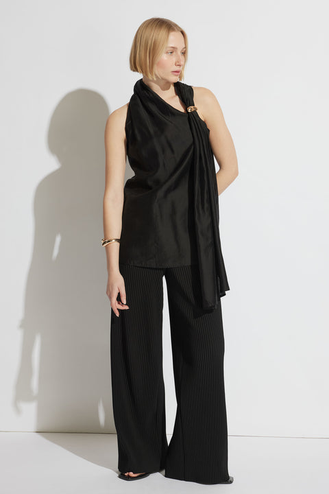 Woman wearing a black draped top with a gold ring detail, paired with wide-leg pleated black pants and gold bracelet.