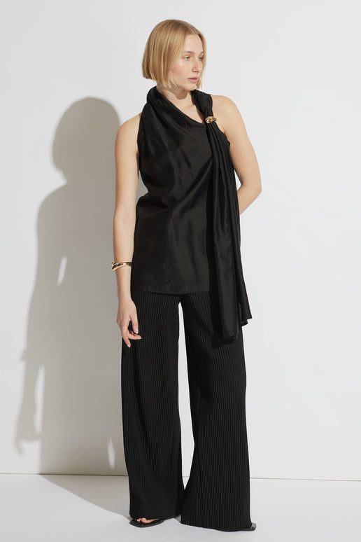 Woman wearing a black draped top with a gold ring detail, paired with wide-leg pleated black pants and gold bracelet.