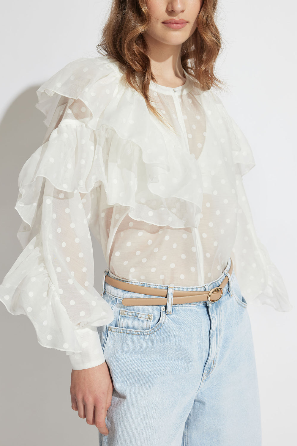 Woman wearing a sheer white spot print blouse with layered ruffles paired with light wash high-waisted jeans