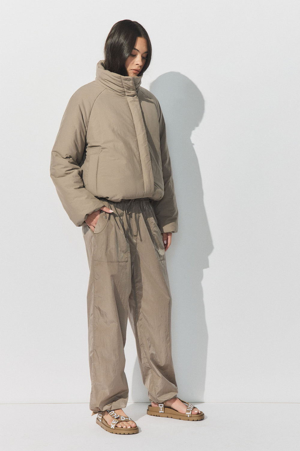 Woman wearing a high-neck padded jacket with relaxed fit drawstring pants and flat strappy sandals against a white wall