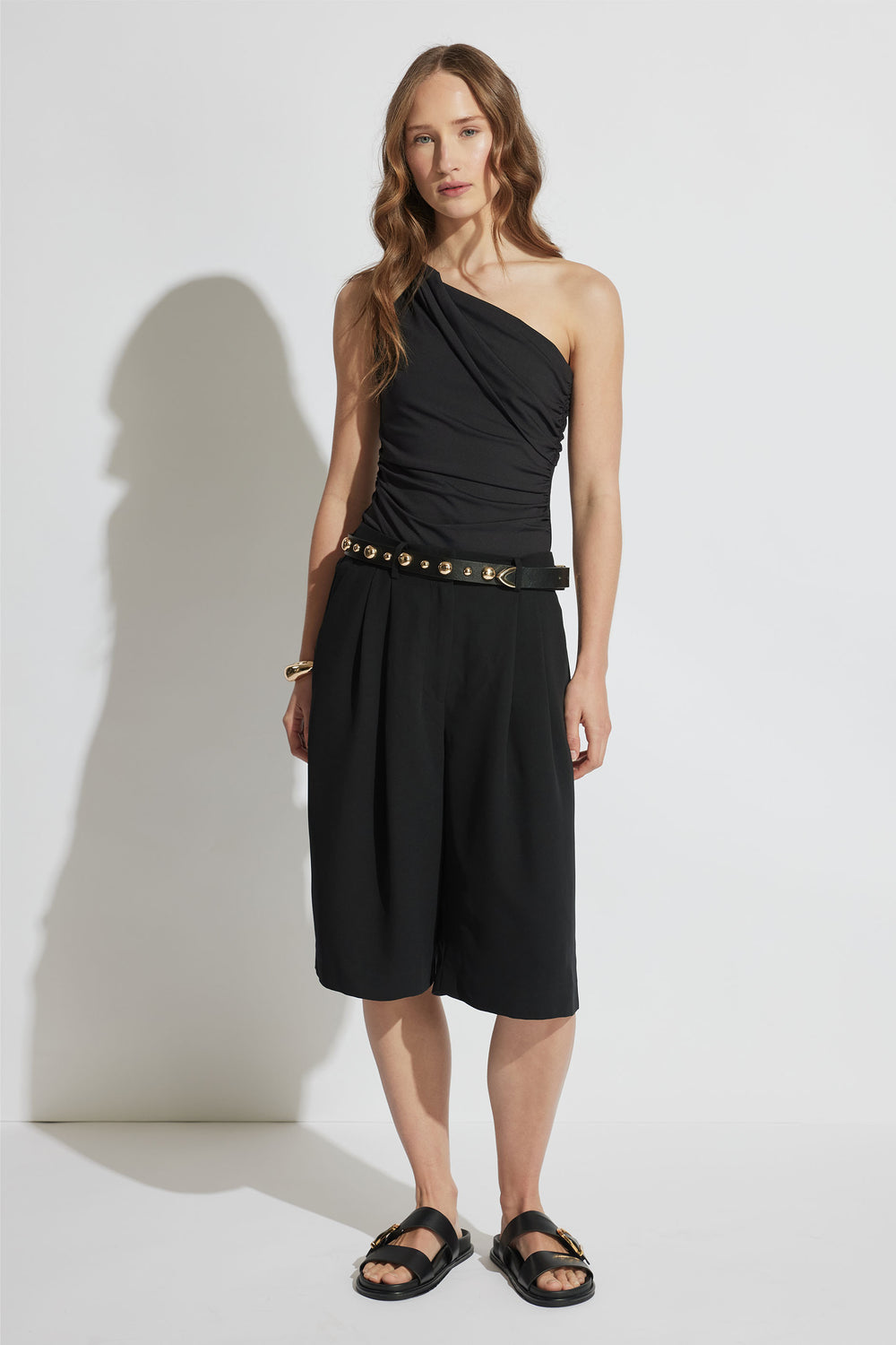 Woman wearing a fitted black one-shoulder twist top paired with wide-leg black culottes and studded belt