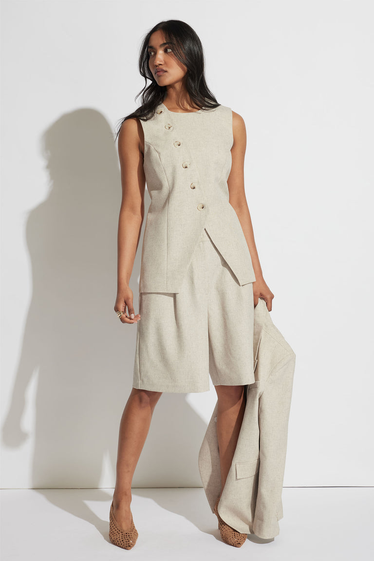 Woman wearing a beige sleeveless longline vest with asymmetrical buttons, styled with matching tailored shorts