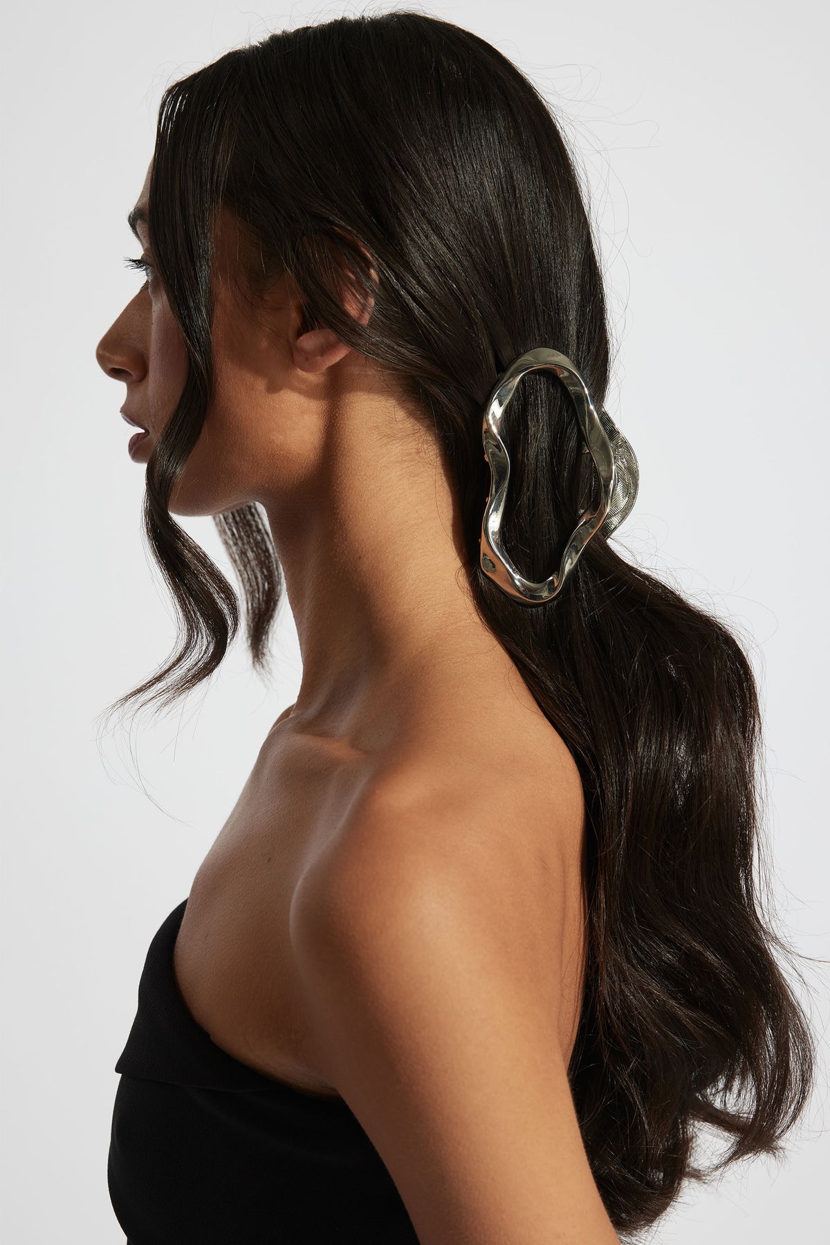 Side profile of woman with long dark hair secured by a silver wavy hair claw, wearing a black strapless top
