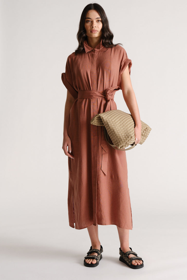 Woman wearing a rust-colored linen blend midi dress with rolled sleeves and a waist tie, accessorized with a woven clutch and sandals