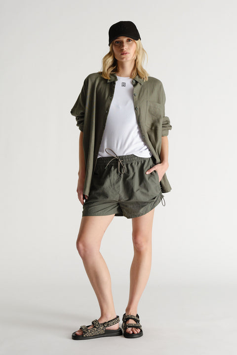 Woman wearing relaxed fit dark green shorts with drawstring waist, paired with a loose shirt, white tee, black cap, and sandals.