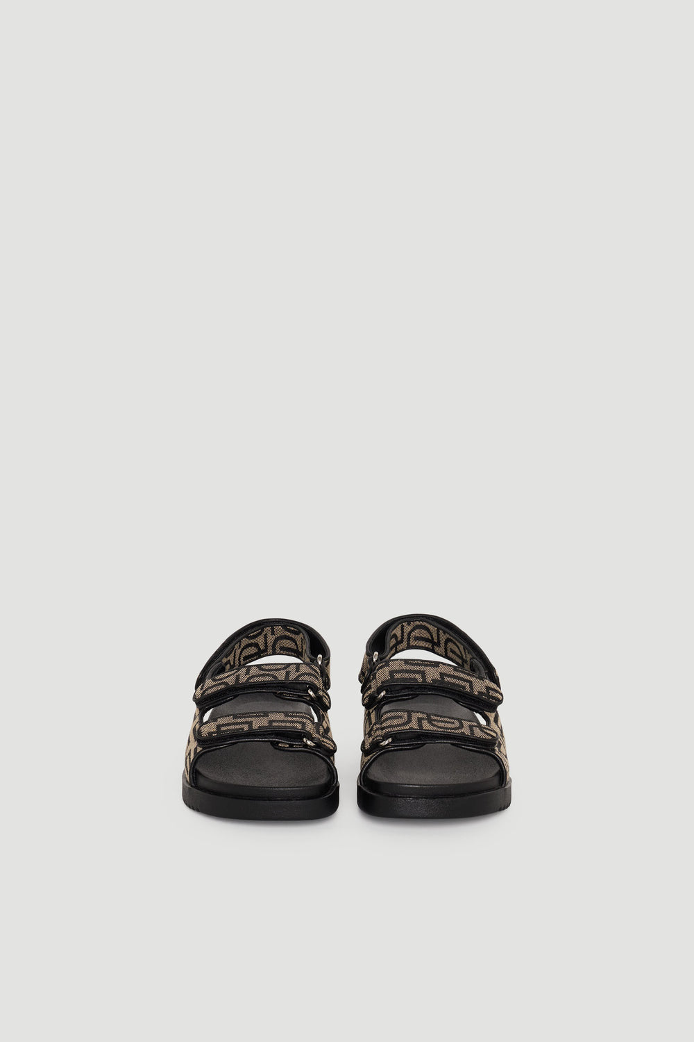 Pair of sandals with black soles and brown monogram straps featuring adjustable buckles, shown front-facing.