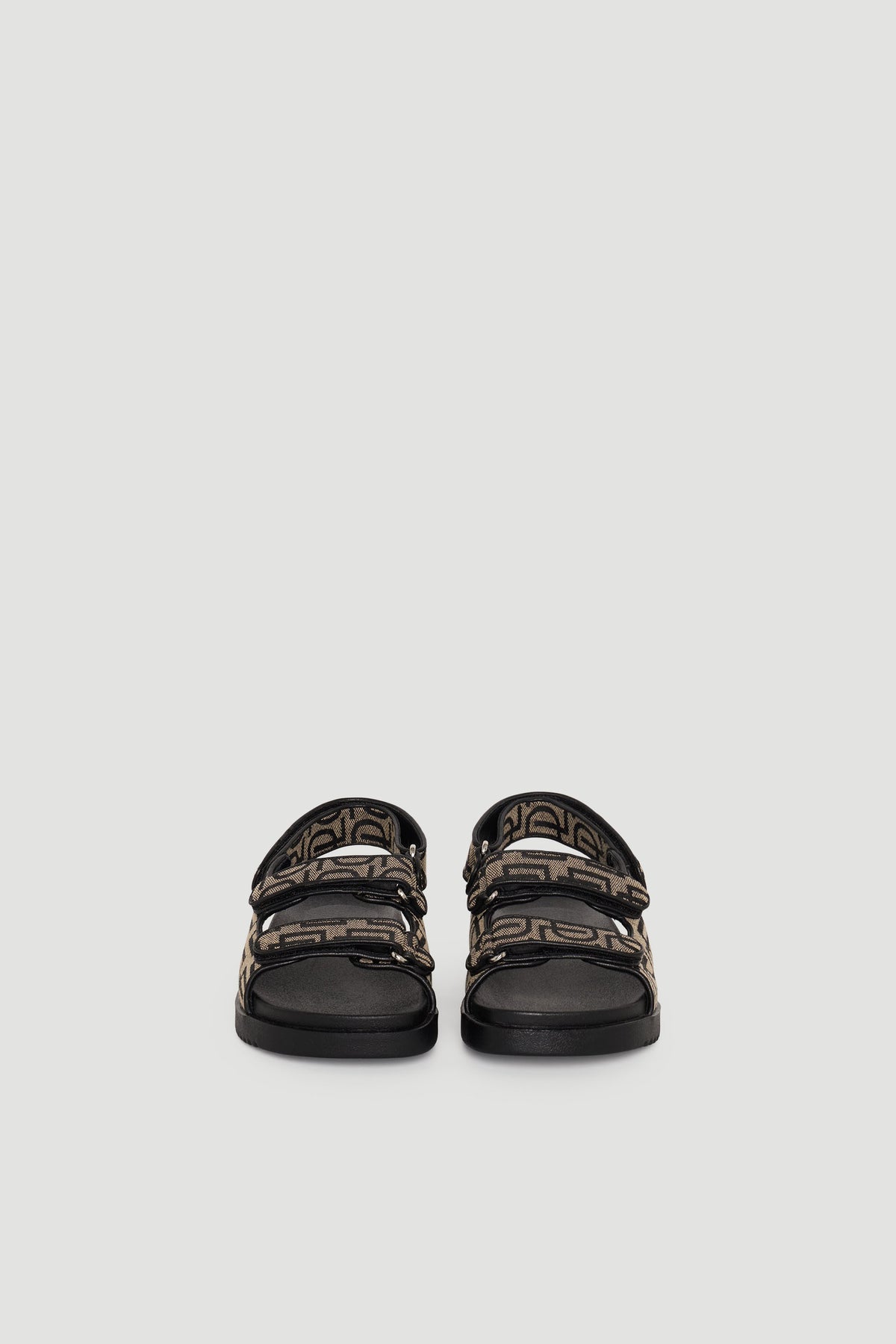 Pair of sandals with black soles and brown monogram straps featuring adjustable buckles, shown front-facing.
