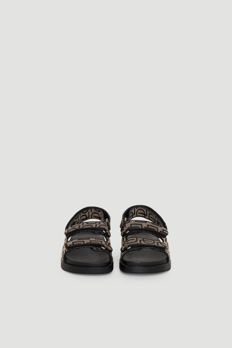 Pair of sandals with black soles and brown monogram straps featuring adjustable buckles, shown front-facing.
