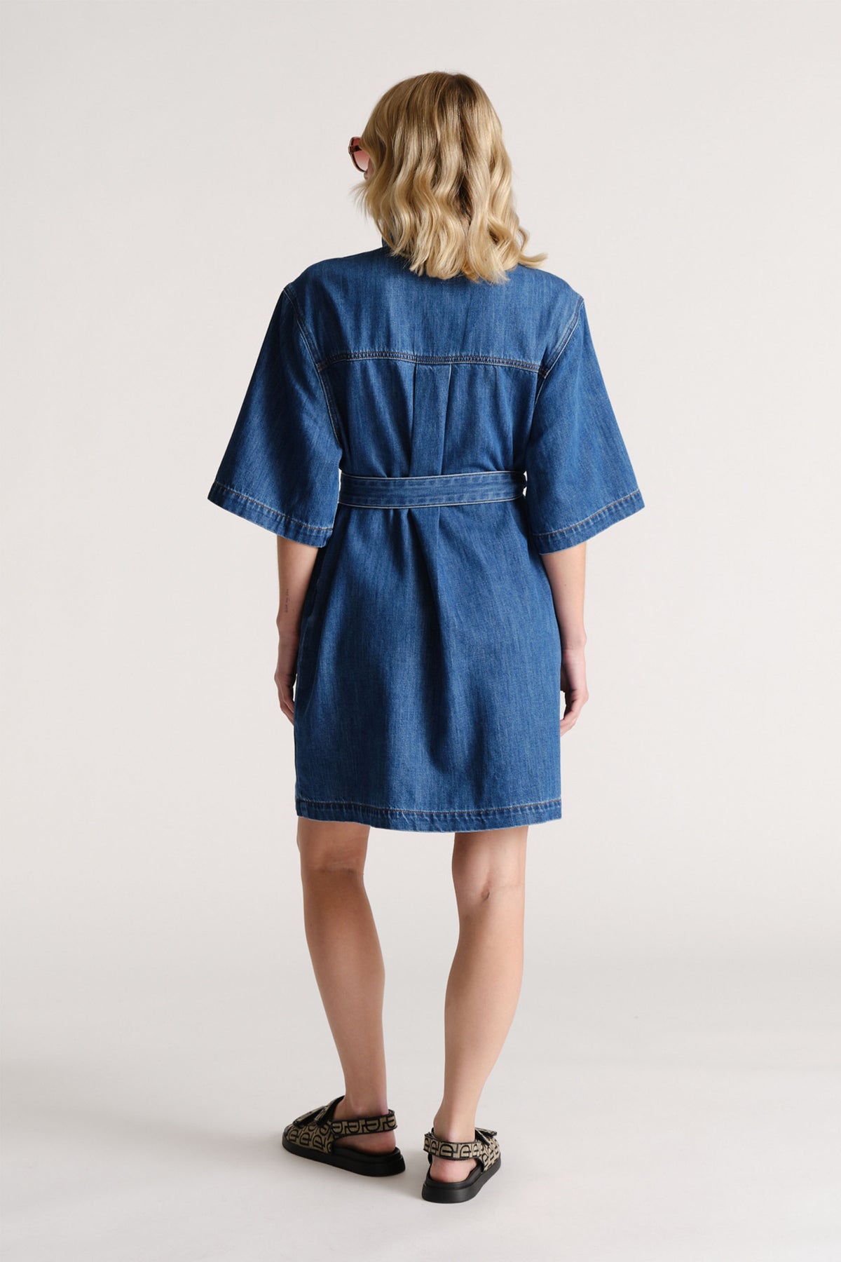Woman wearing a mid-length blue denim dress with wide sleeves and a belted waist, paired with black sandals.