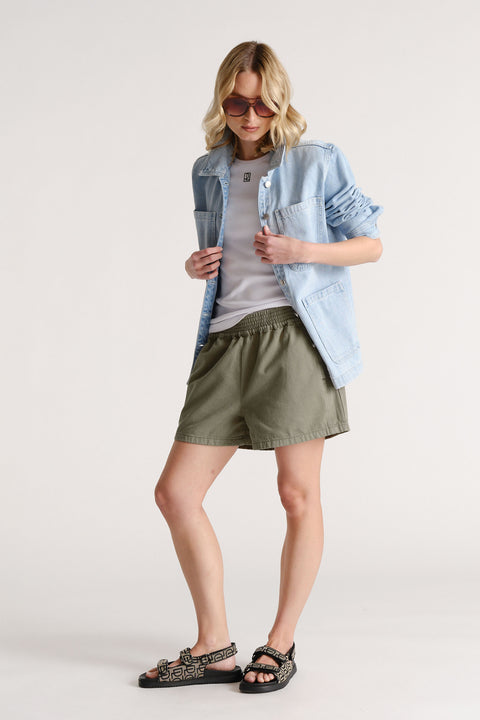 Woman wearing olive green pull-on shorts paired with a white tee, light denim jacket, and patterned sandals.