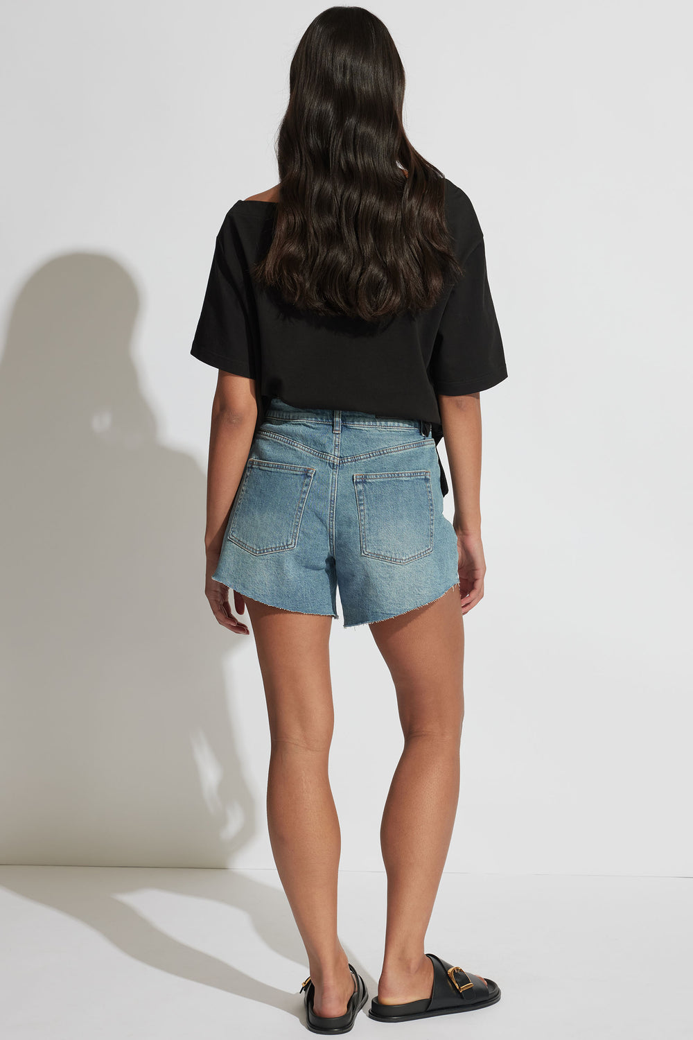 Woman wearing light blue denim shorts with raw hem edges, paired with a loose black top and black slip-on shoes.