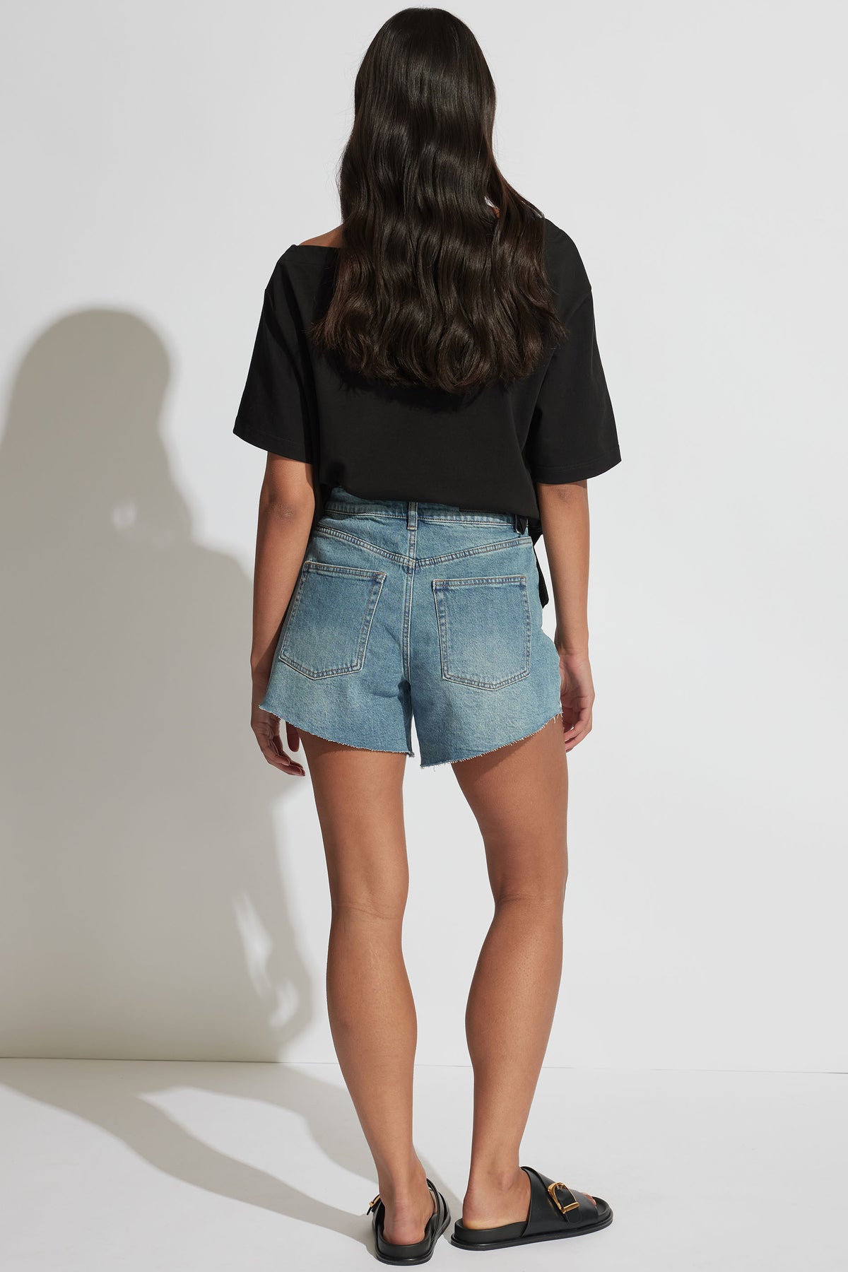 Woman wearing light blue denim shorts with raw hem edges, paired with a loose black top and black slip-on shoes.