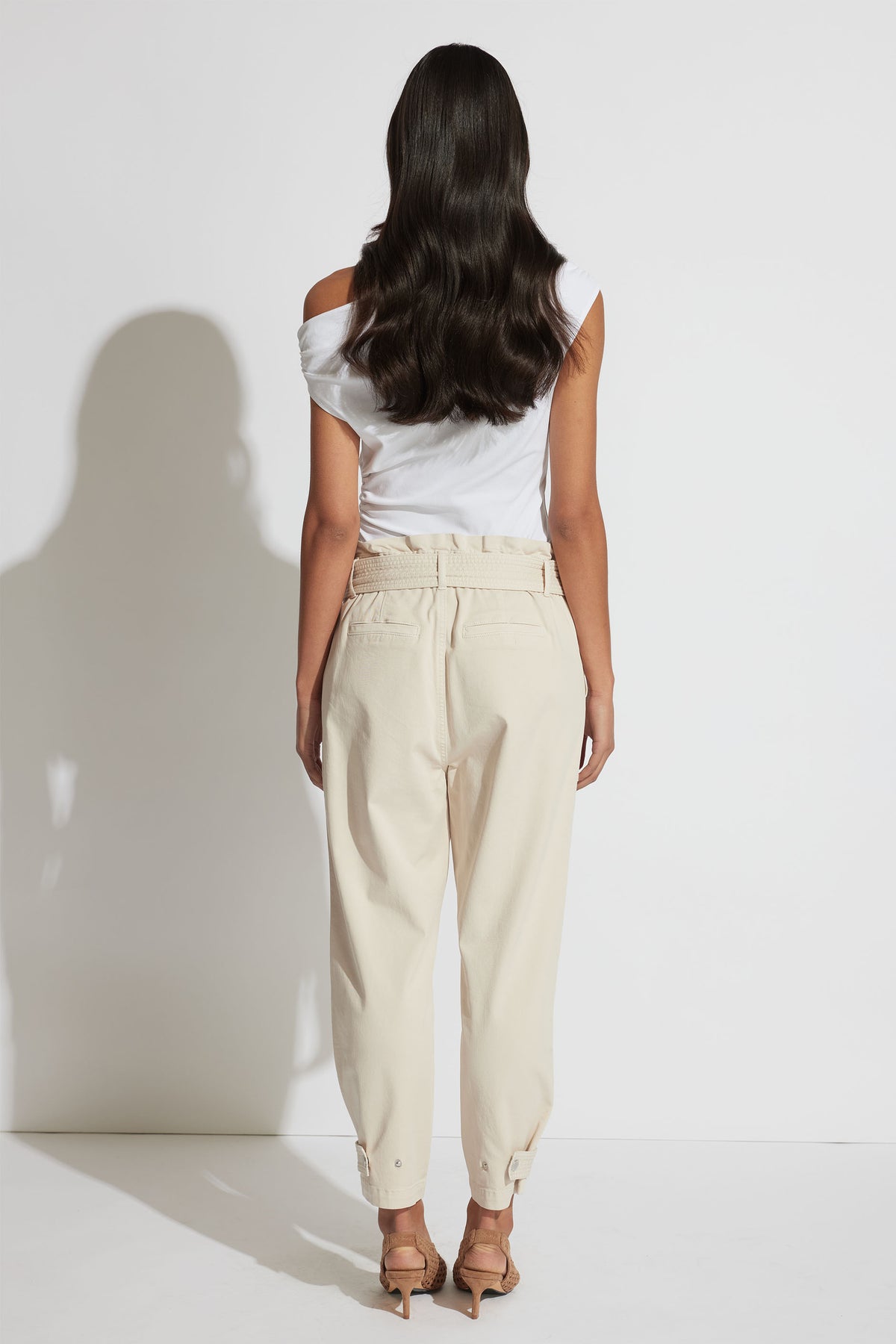 Woman wearing high-waisted beige pull-on pants with ankle straps paired with a white off-shoulder top and heels.