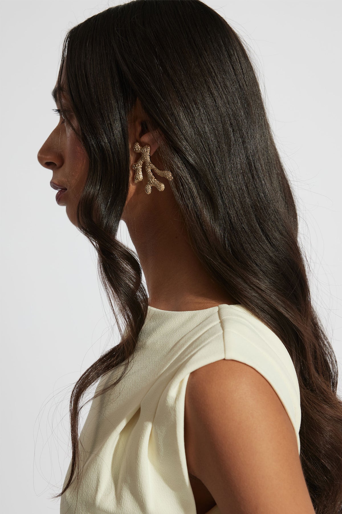 Profile view of woman with long dark hair wearing textured gold coral-shaped statement earrings and a cream sleeveless top