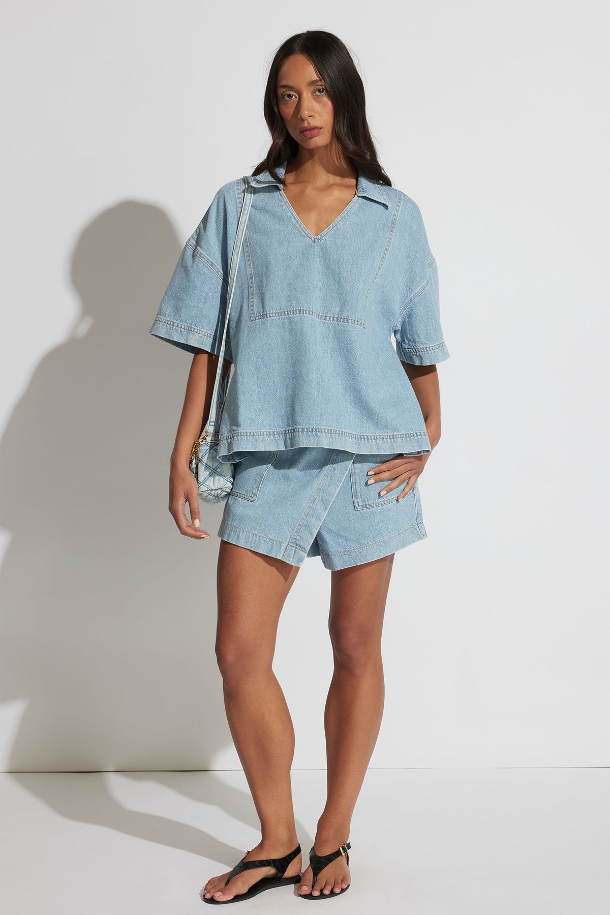 Woman wearing a light wash oversized denim shirt with matching high-waisted shorts and black sandals, posing front view.