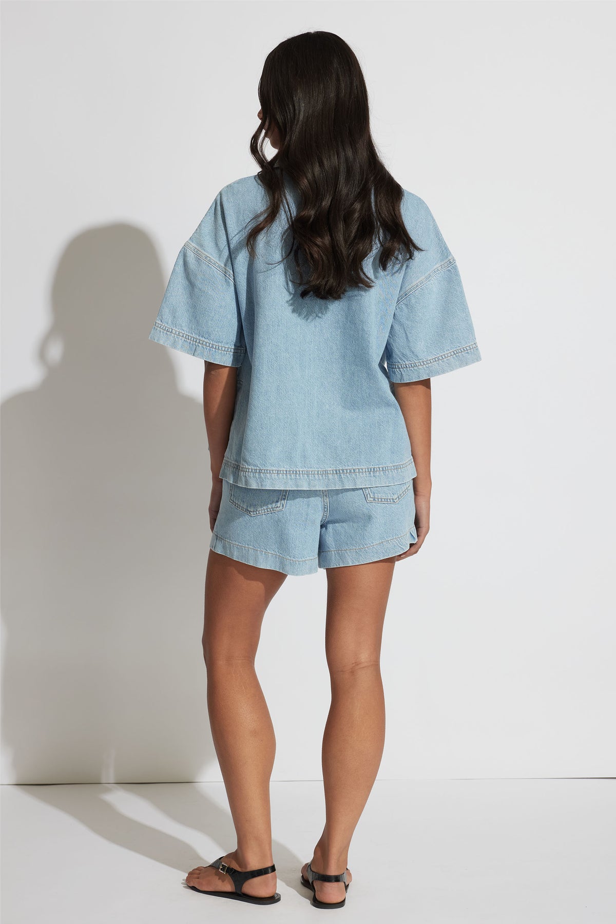 Woman wearing a relaxed fit light blue denim shirt and matching shorts, paired with black sandals, shown from behind.