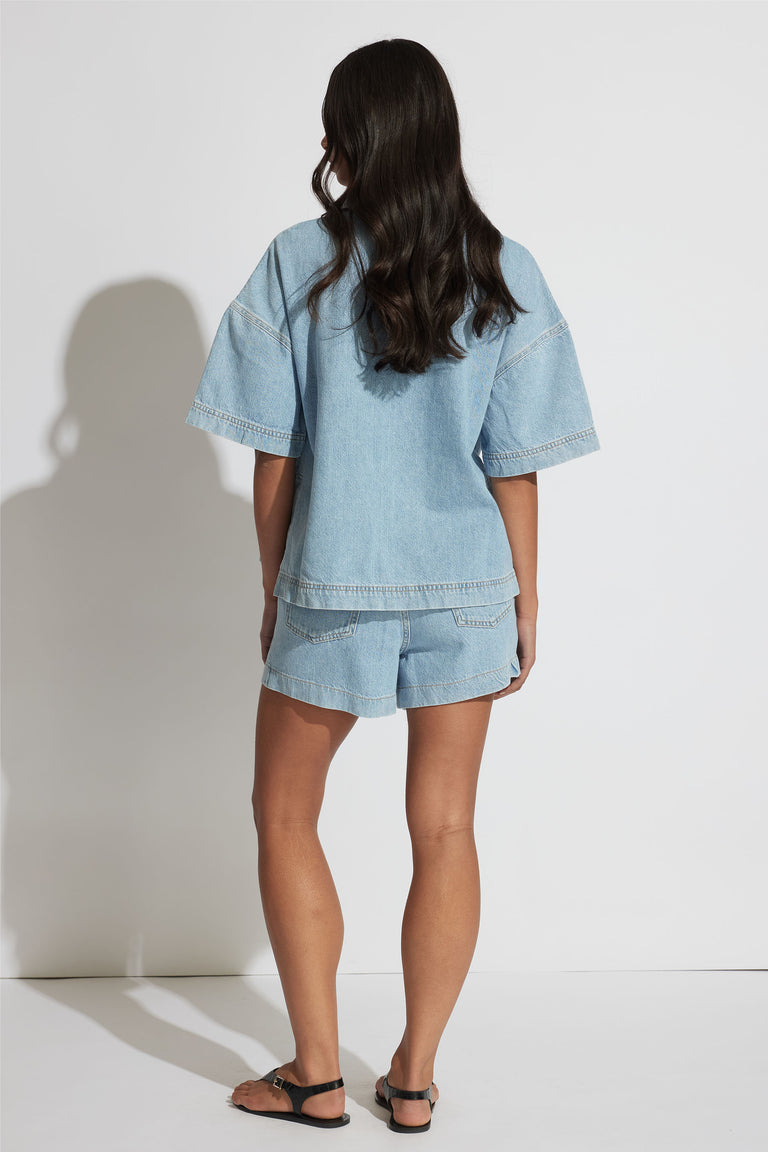 Woman wearing a relaxed fit light blue denim shirt and matching shorts, paired with black sandals, shown from behind.