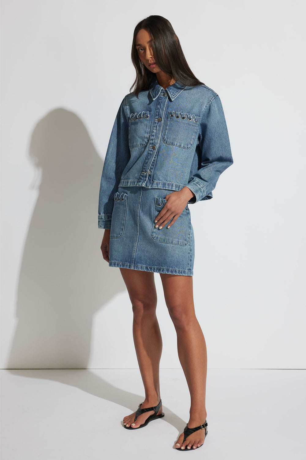 Woman wearing a medium blue denim jacket and matching skirt set styled with black thong sandals, standing against a white wall.