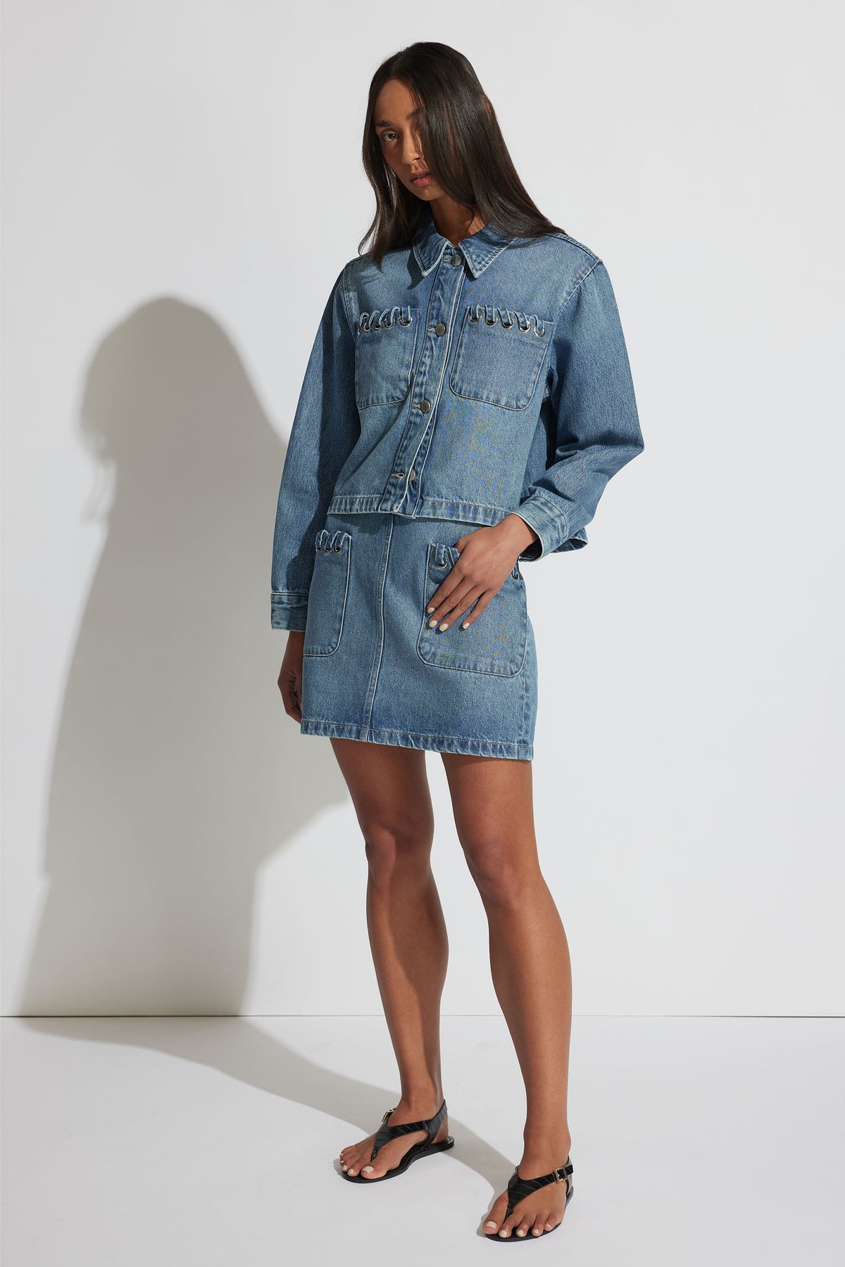 Woman wearing a medium blue denim jacket and matching skirt set styled with black thong sandals, standing against a white wall.