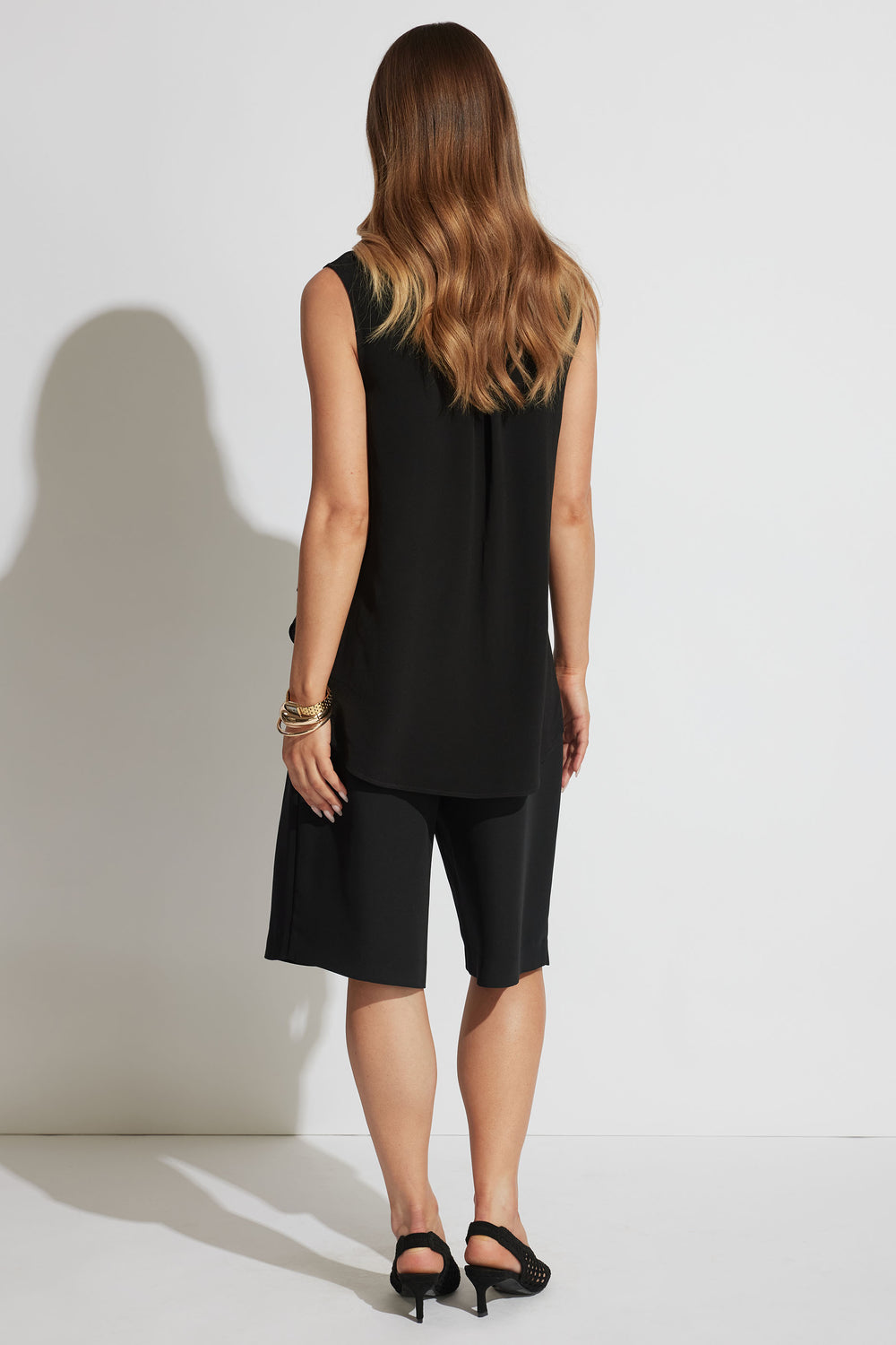 Woman wearing a loose black woven tank paired with knee-length black shorts and heeled sandals in a minimalist look.