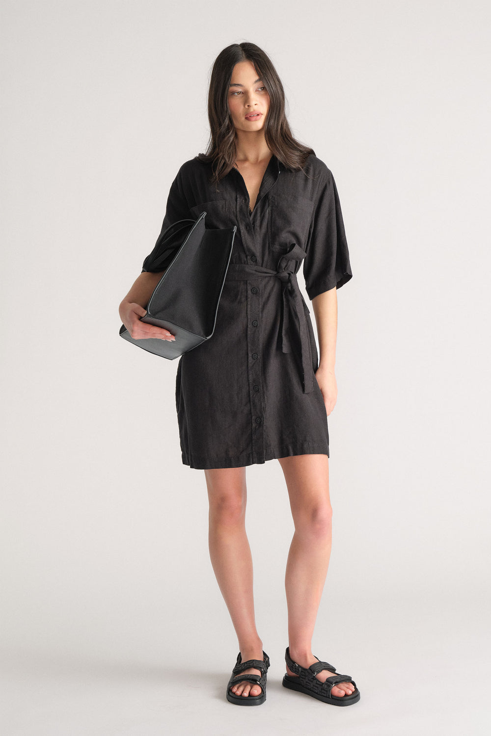 Woman wearing a black linen blend short dress with a waist tie, styled with black sandals and a large black tote bag.