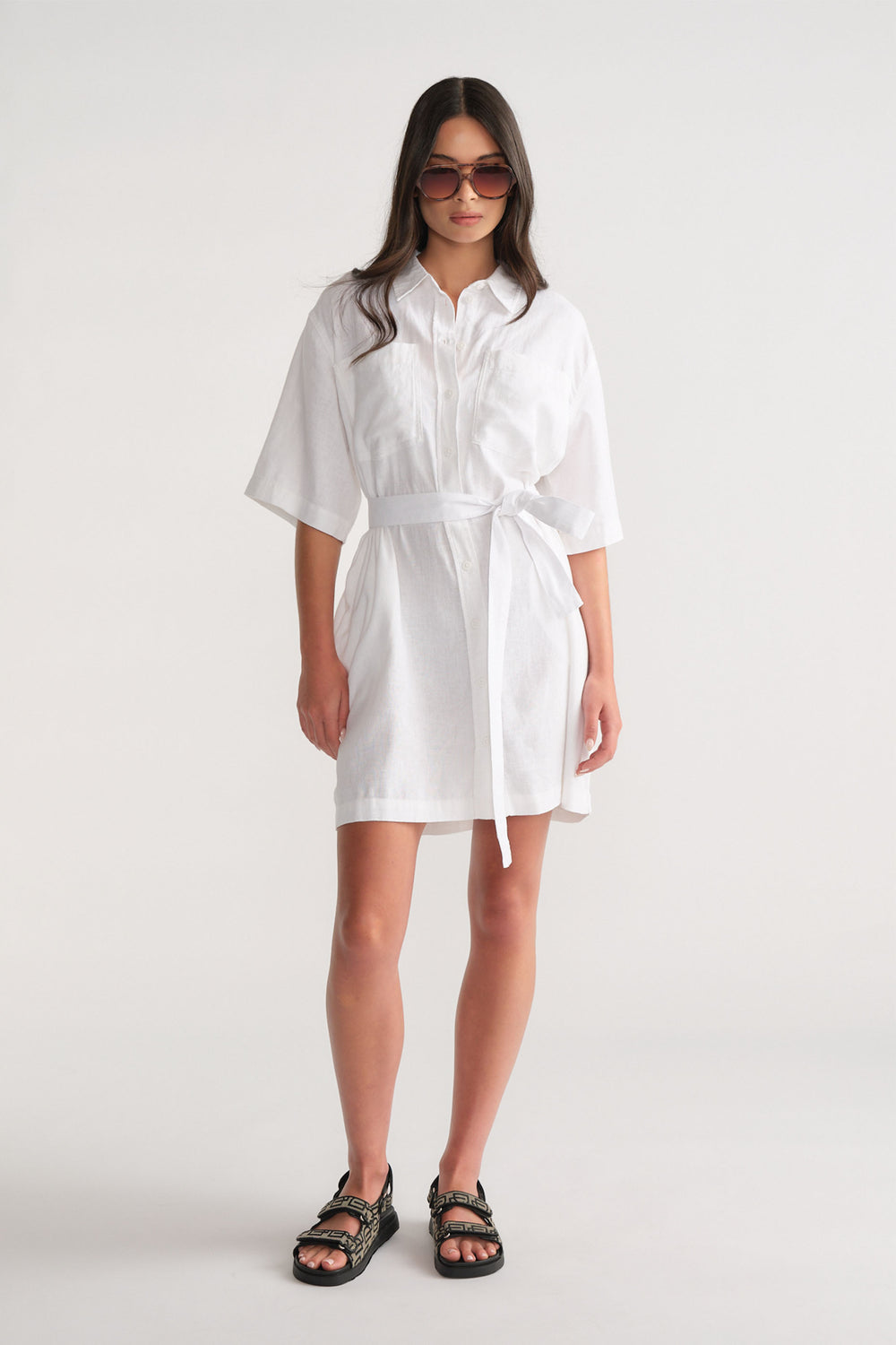 Woman wearing a white linen blend short dress with a self-tie waist, styled with black sandals and oversized sunglasses.