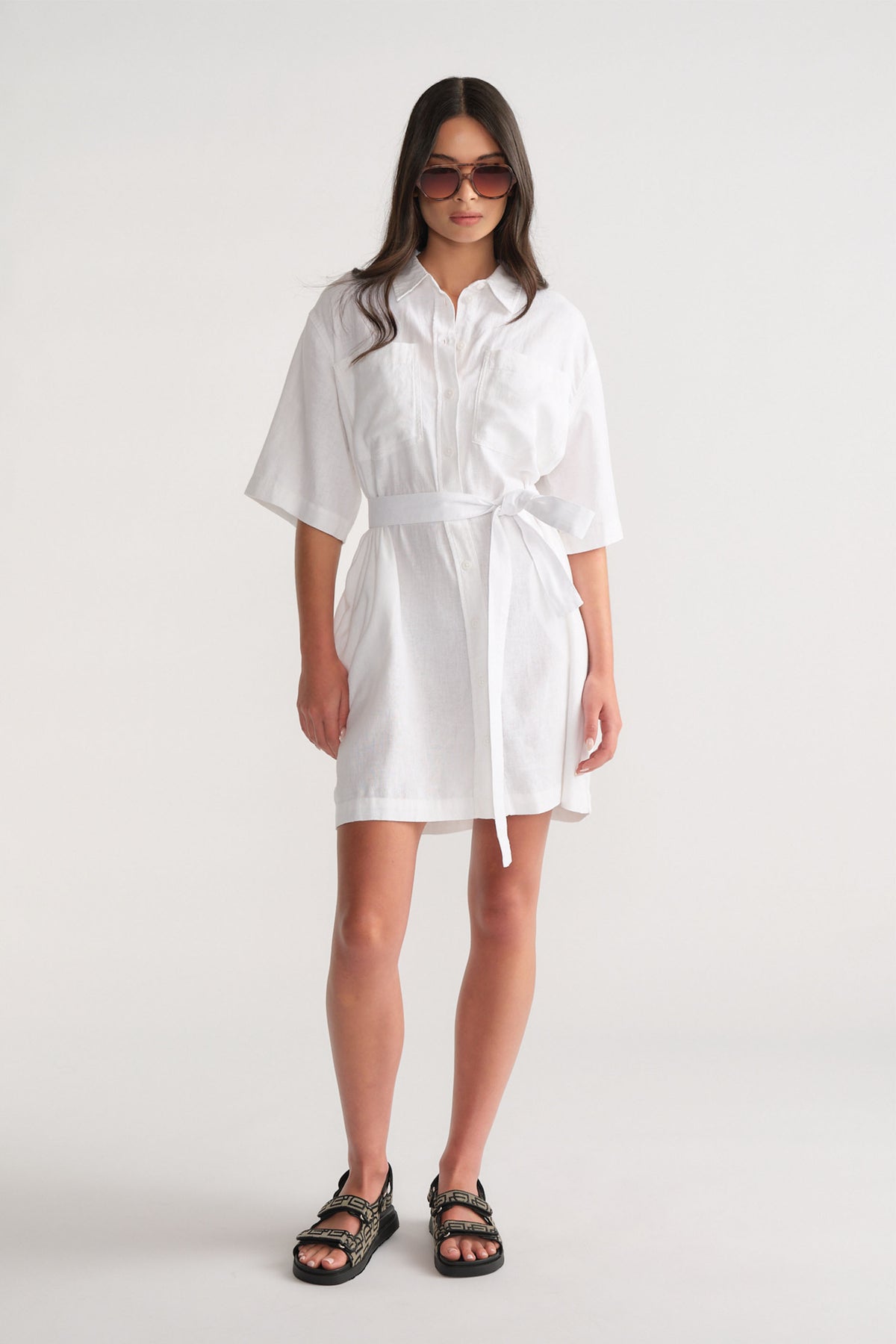 Woman wearing a white linen blend short dress with a self-tie waist, styled with black sandals and oversized sunglasses.