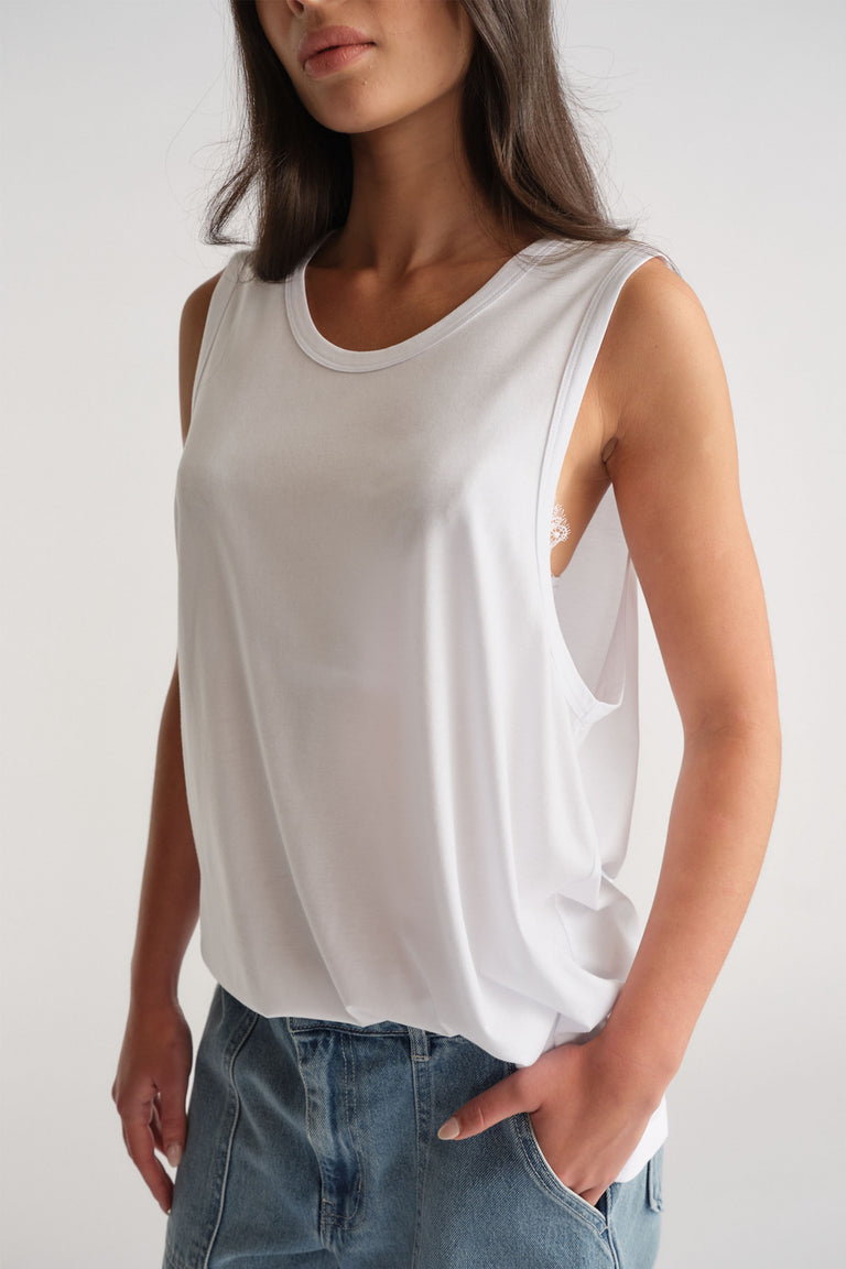 Woman wearing a relaxed white sleeveless tank with wide armholes paired with casual blue denim jeans.