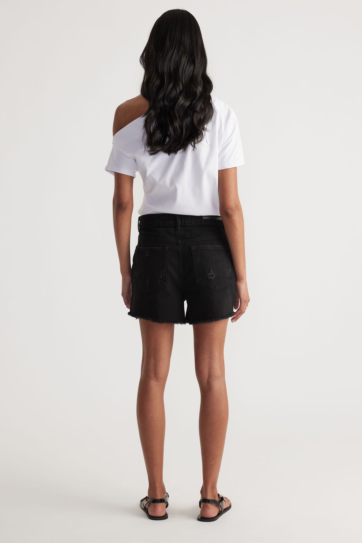 Woman seen from behind wearing a white off-shoulder top paired with black distressed denim shorts and sandals