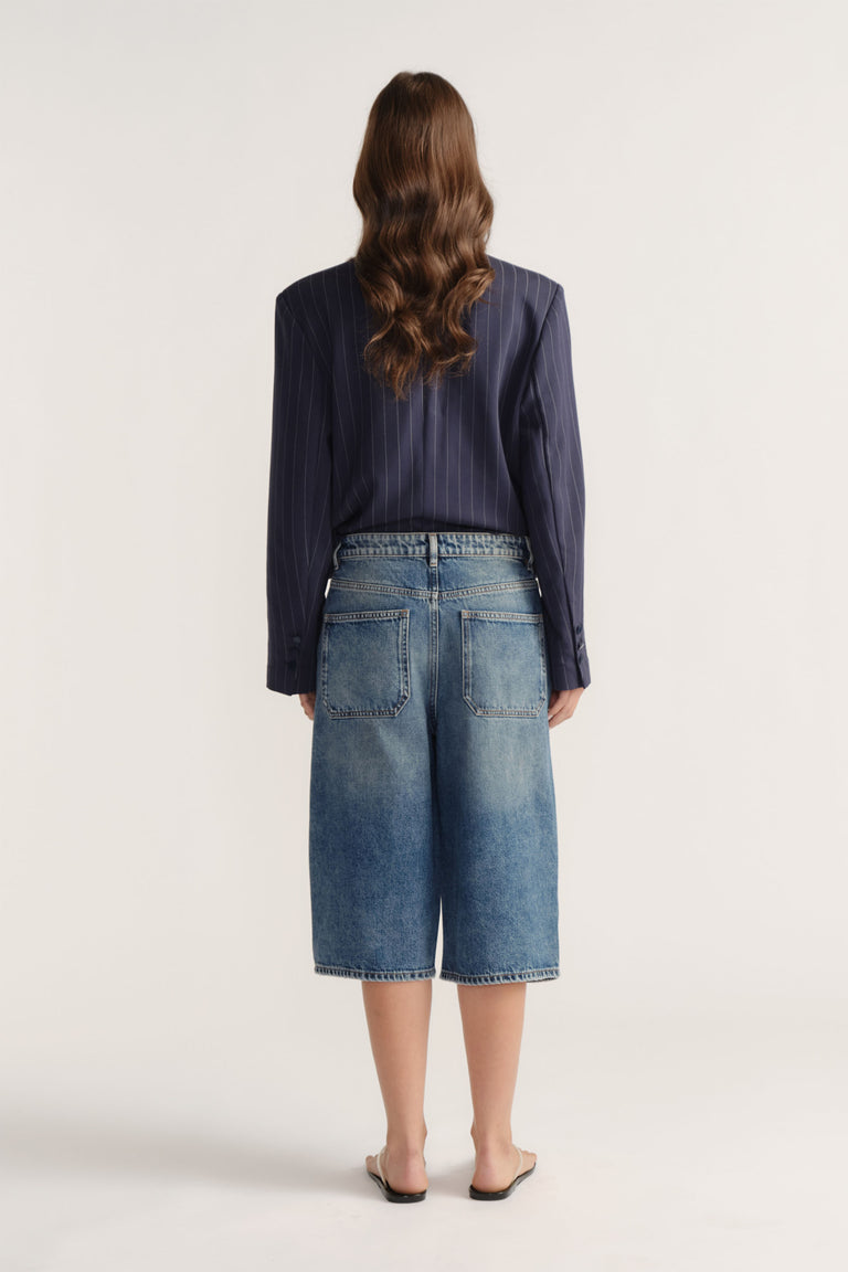 Rear view of D-Luxe Denim Long Line Short in mid-blue cotton denim styled with a navy pinstripe blazer by Decjuba Australian brand