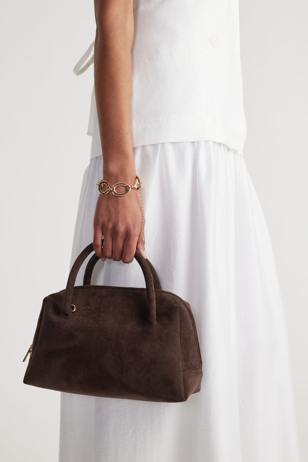 Woman wearing a gold open chain bracelet paired with a white outfit holding a dark brown suede handbag