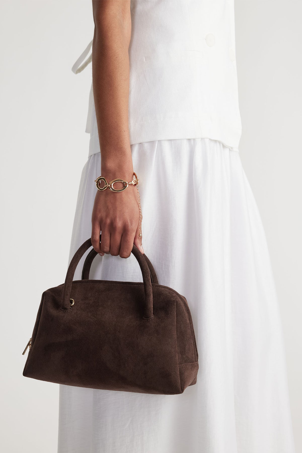 Woman wearing a gold open chain bracelet paired with a white outfit holding a dark brown suede handbag