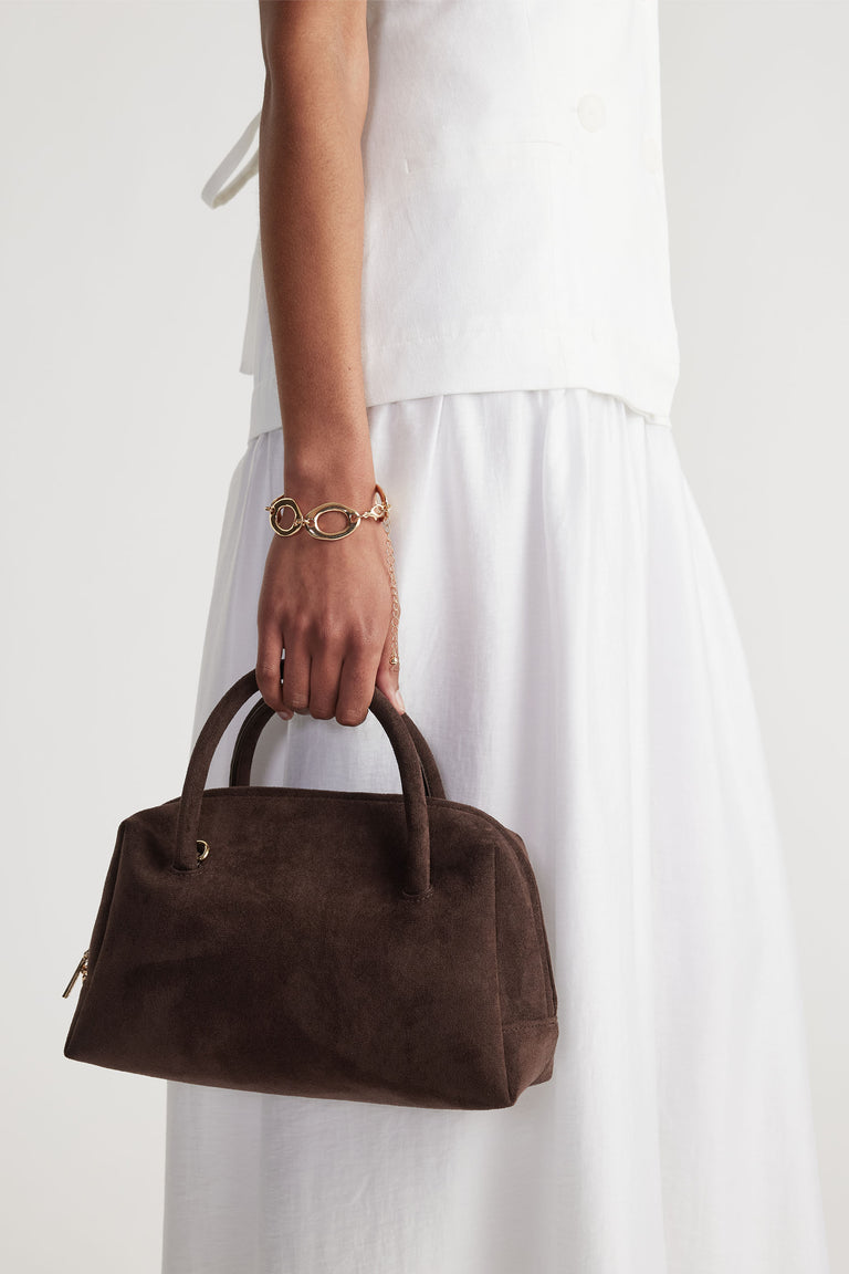 Woman wearing a gold open chain bracelet paired with a white outfit holding a dark brown suede handbag