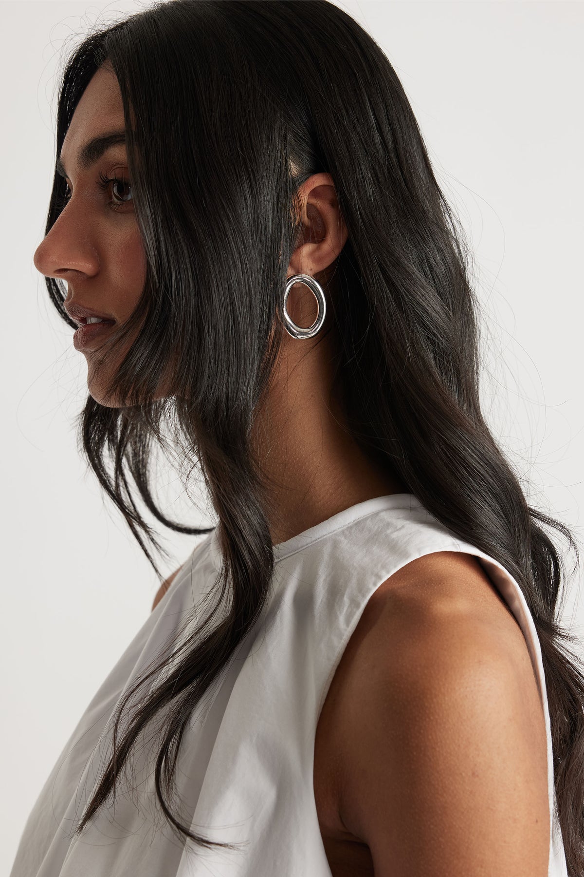 Side profile of woman wearing modern silver open hoop earrings with dark hair and white sleeveless top