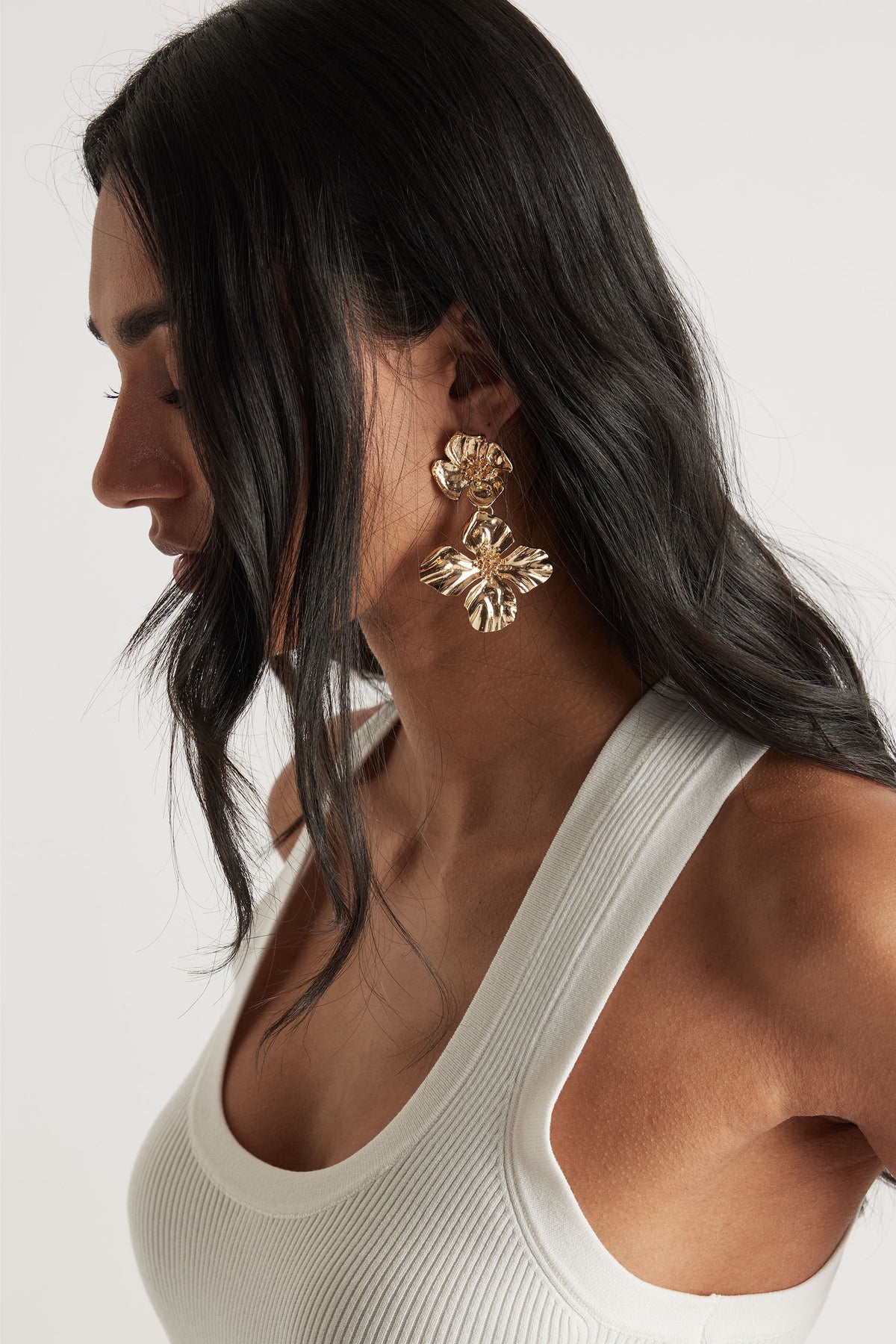 Side profile of woman wearing textured gold floral drop earrings paired with a white ribbed tank top