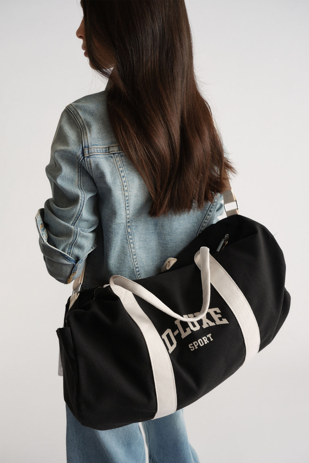 Woman wearing a light denim jacket holding a black duffle bag with white handles for a casual, sporty look