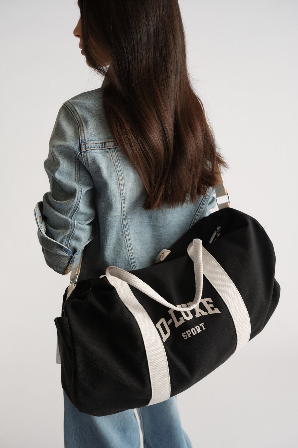 Woman wearing a light denim jacket holding a black duffle bag with white handles for a casual, sporty look