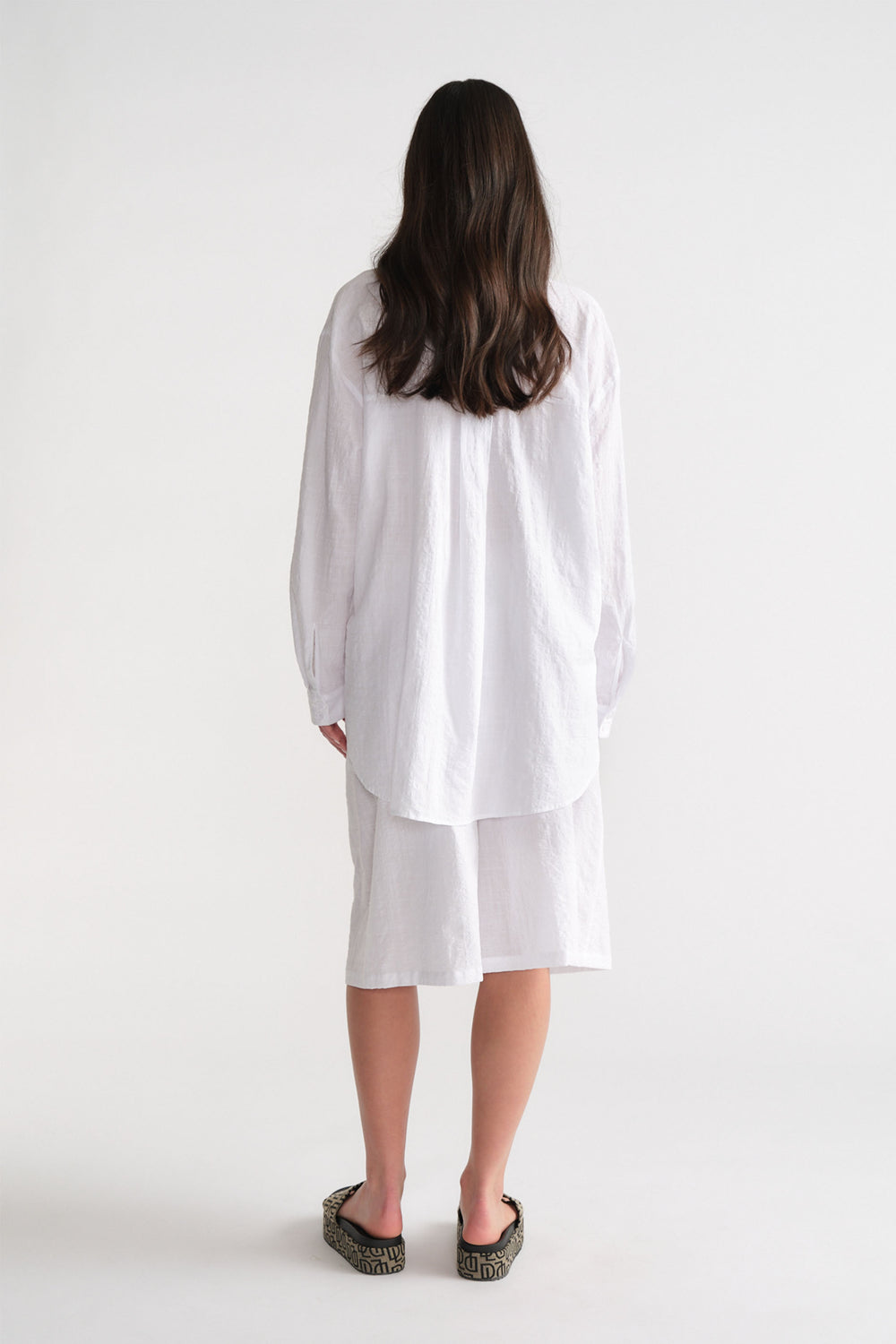 Woman seen from behind wearing a white long-sleeve shirt and relaxed white Bermuda shorts with patterned slip-on shoes