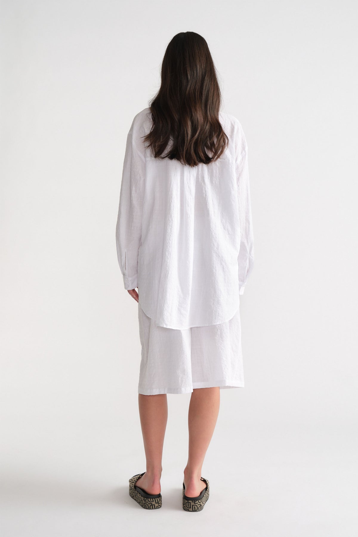 Woman seen from behind wearing a white long-sleeve shirt and relaxed white Bermuda shorts with patterned slip-on shoes