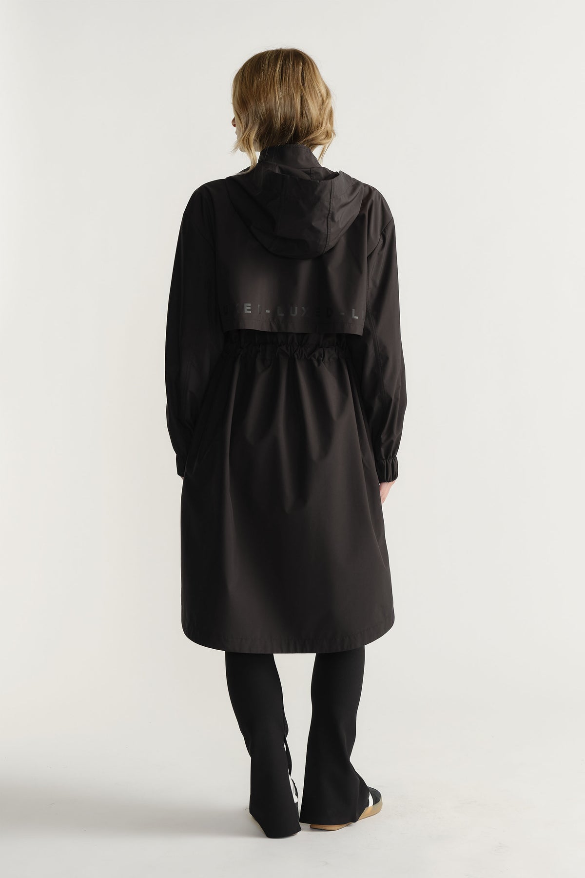 Back view of woman wearing black D-Luxe Rain Jacket with hood and cinched waist styled with black pants by Decjuba