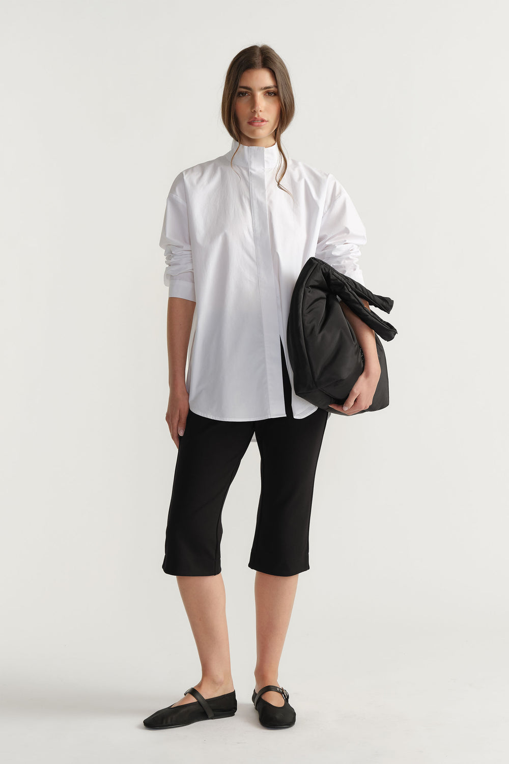 Woman wearing black cropped pants paired with a crisp white long-sleeve shirt and black flats, holding a black jacket.