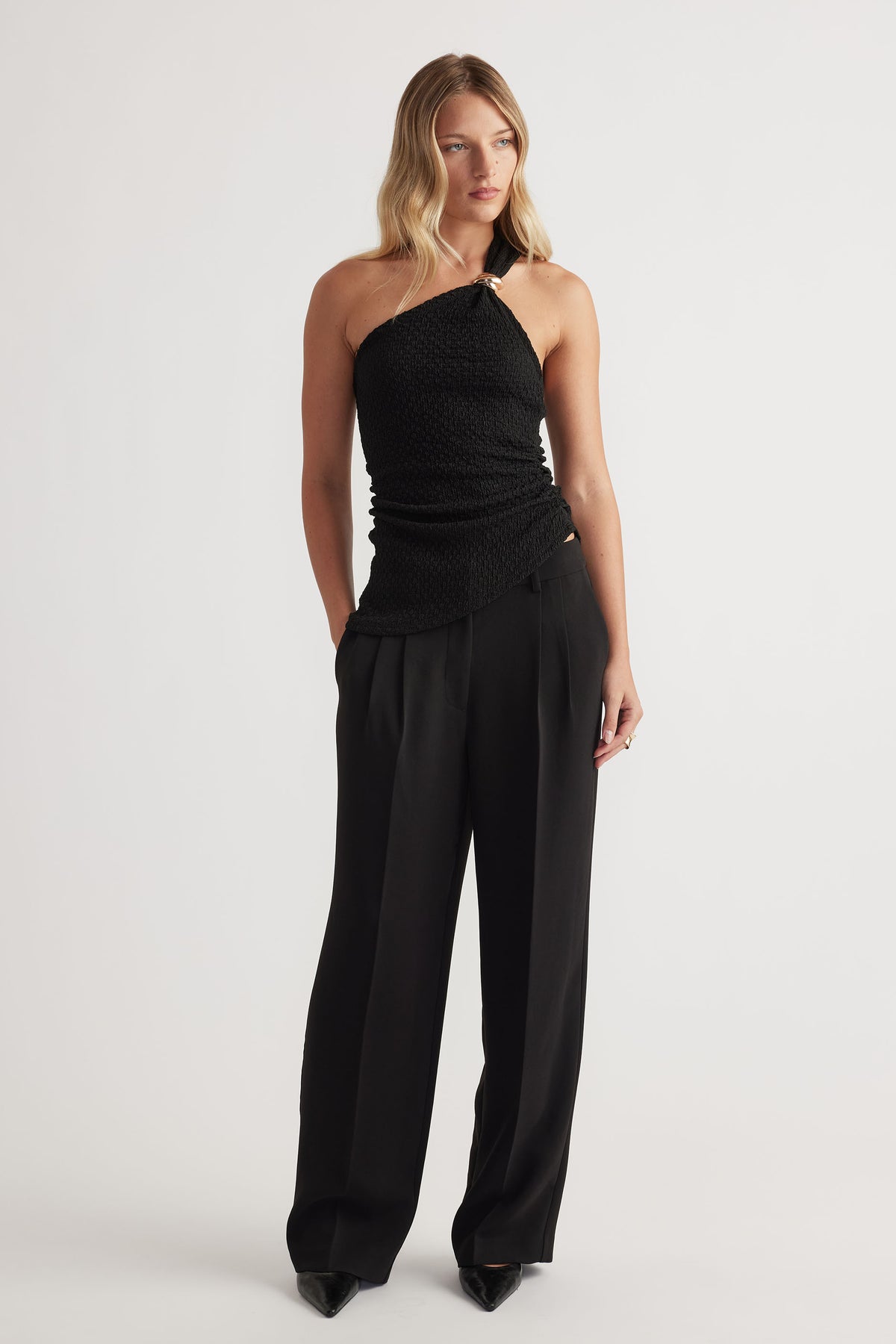 Woman wearing a textured black one-shoulder tank paired with high-waisted wide-leg black trousers and pointed heels