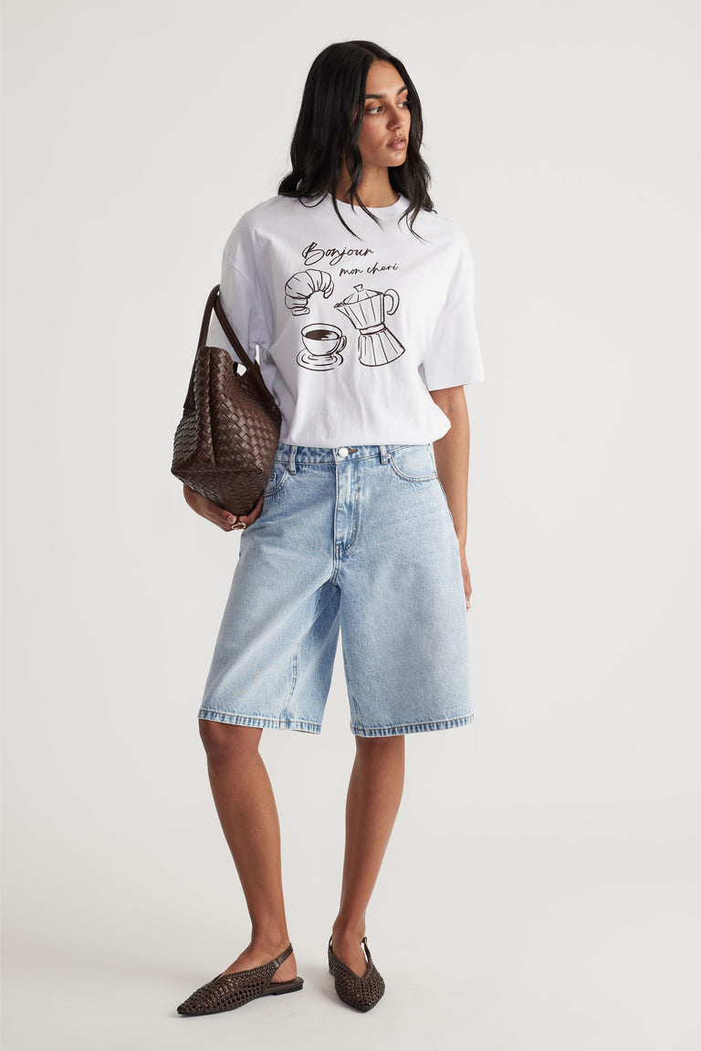 Woman wearing a relaxed white printed tee with light blue long denim shorts and brown woven bag and flats